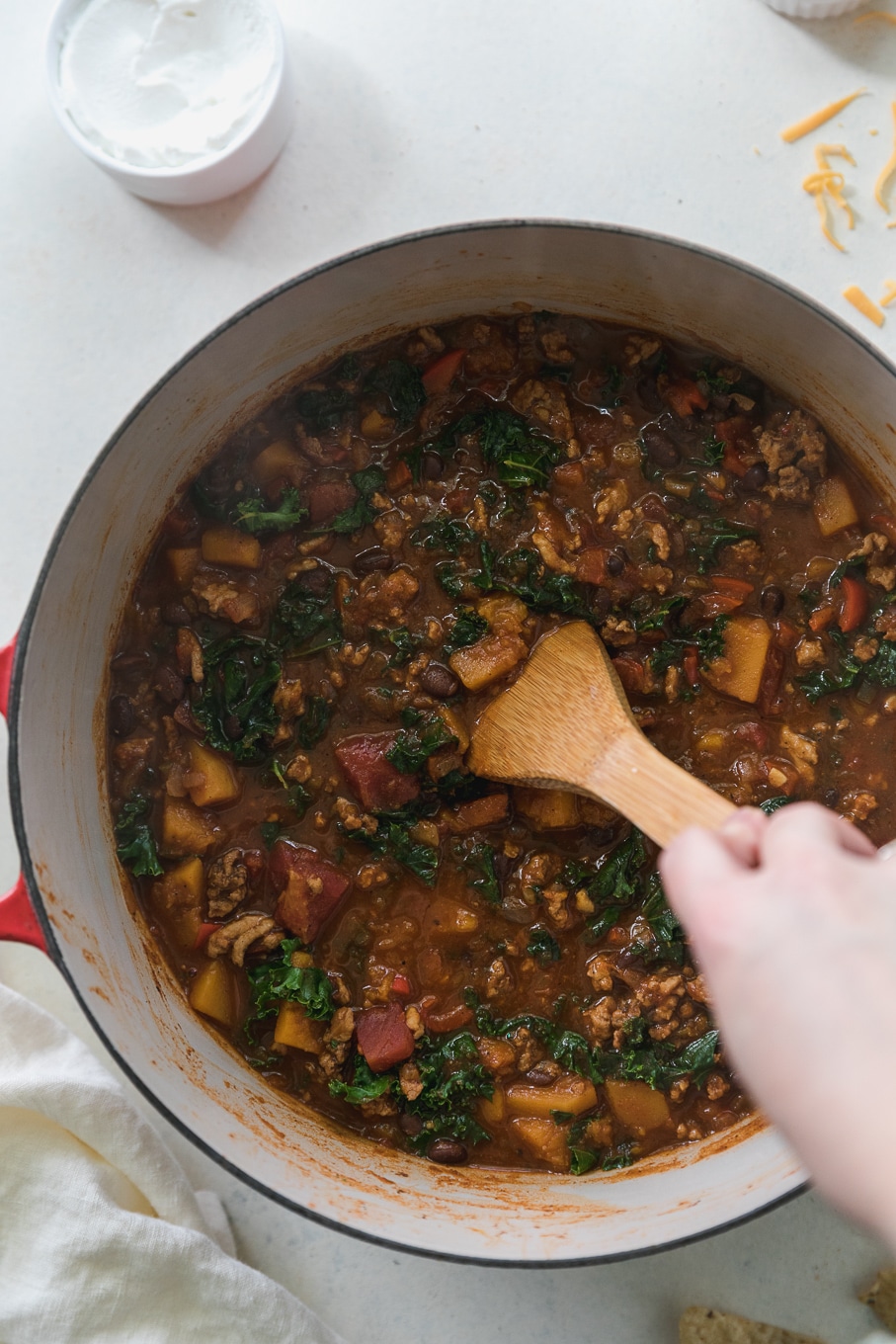 Overhead shot of a pot with red handles filled with chili and a hand stirring it with a wooden spoon