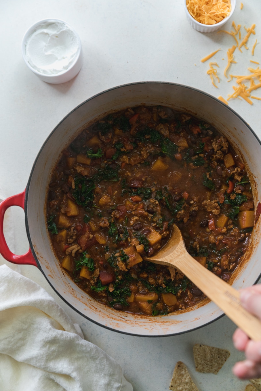 Overhead shot of a pot with red handles filled with chili and a hand stirring it with a wooden spoon
