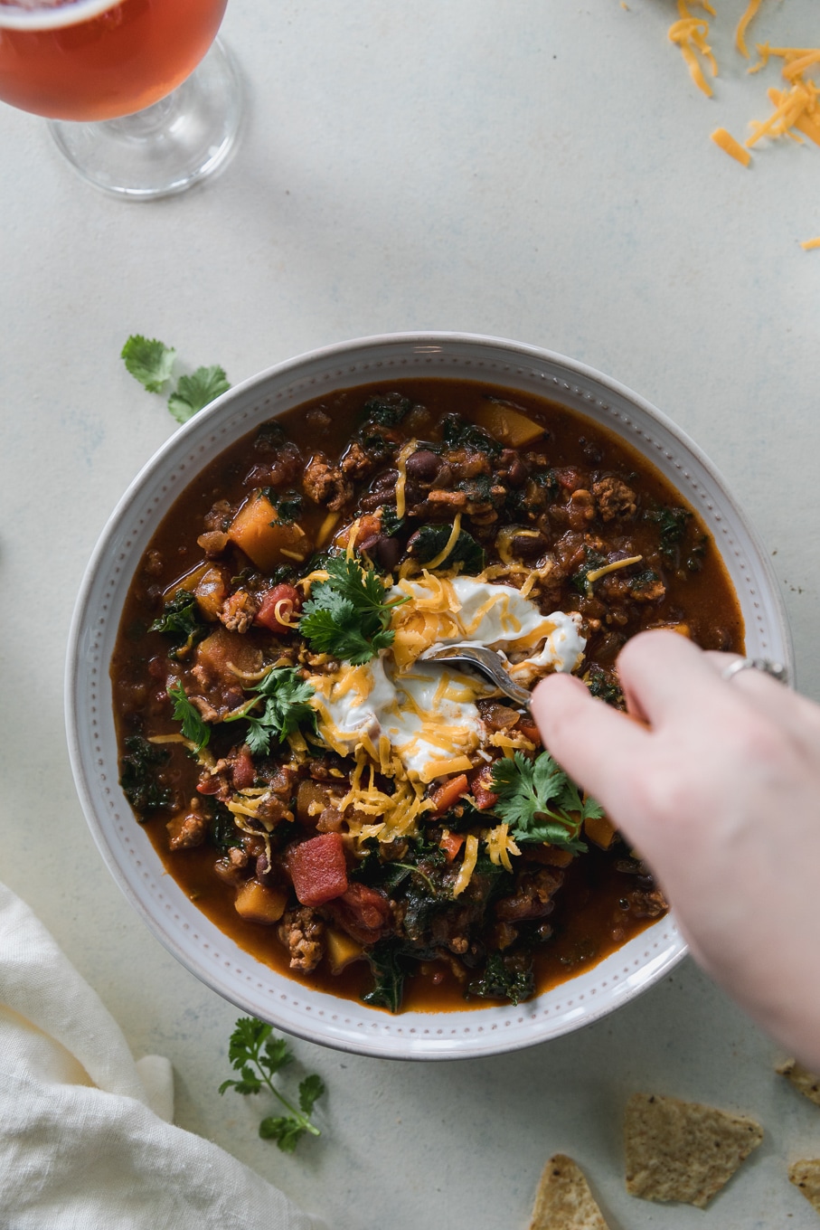 Overhead shot of a bowl of chili topped with sour cream, shredded cheddar, and cilantro, with a hand stirring the chili with a spoon