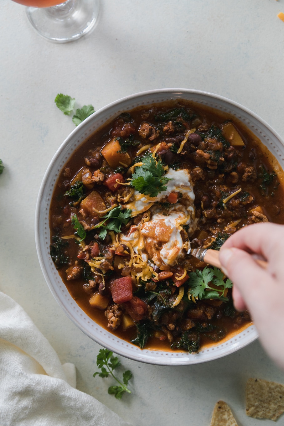 Overhead close up shot of a bowl of chili topped with sour cream, shredded cheddar, and cilantro, with a hand stirring the chili with a spoon