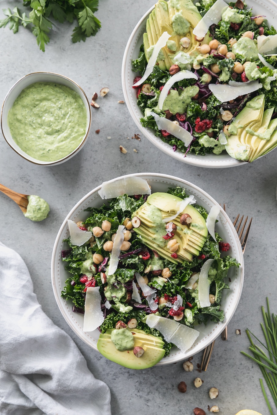 Overhead shot of a kale salad topped with sliced avocado, parmesan shavings, hazelnuts, chickpeas, and dried cranberries