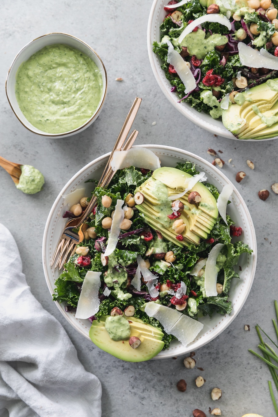 Overhead shot of a kale salad topped with sliced avocado, parmesan shavings, hazelnuts, chickpeas, and dried cranberries with a gold fork resting in the bowl and a bowl of creamy green dressing off to the side