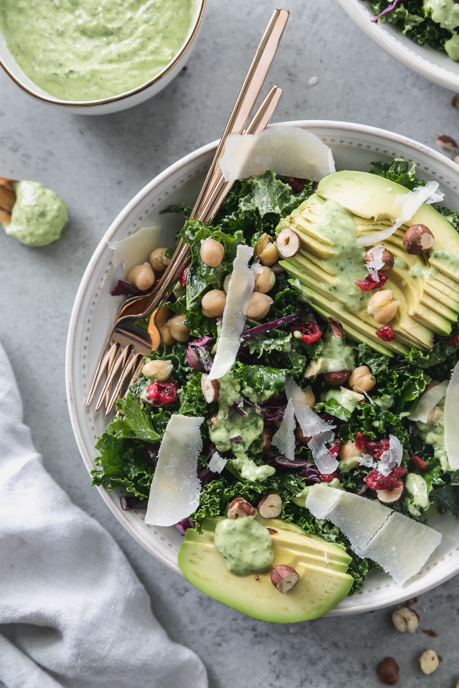 Overhead close up shot of a kale salad topped with sliced avocado, parmesan shavings, hazelnuts, chickpeas, and dried cranberries with a gold fork resting in the bowl