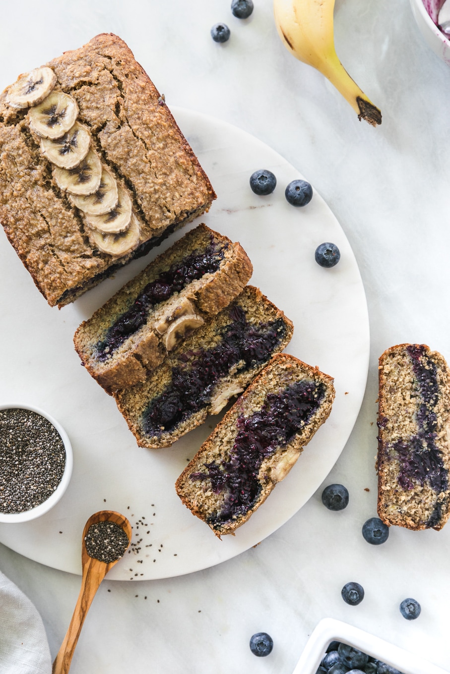 Overhead shot of a loaf of blueberry chia jam stuffed banana bread with 4 slices cut off and a bowl of chia seeds next to it