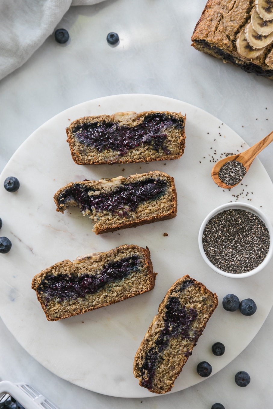 Overhead shot of slices of blueberry chia jam stuffed banana bread with a bite taken out of one of the slices and a bowl of chis seeds with a wooden spoon next to it
