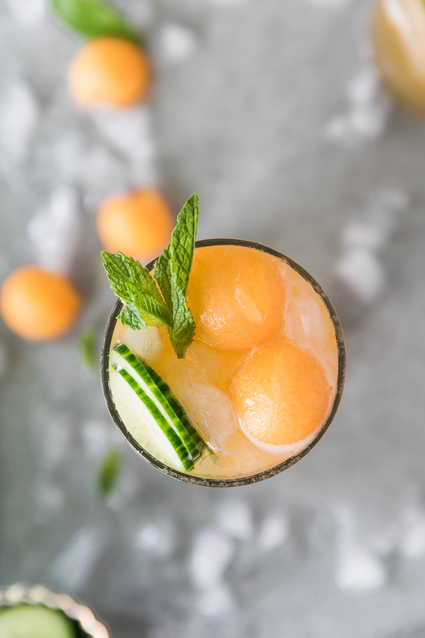 Overhead close up shot of a cocktail topped with melon balls, sliced cucumber, and a spring of mint