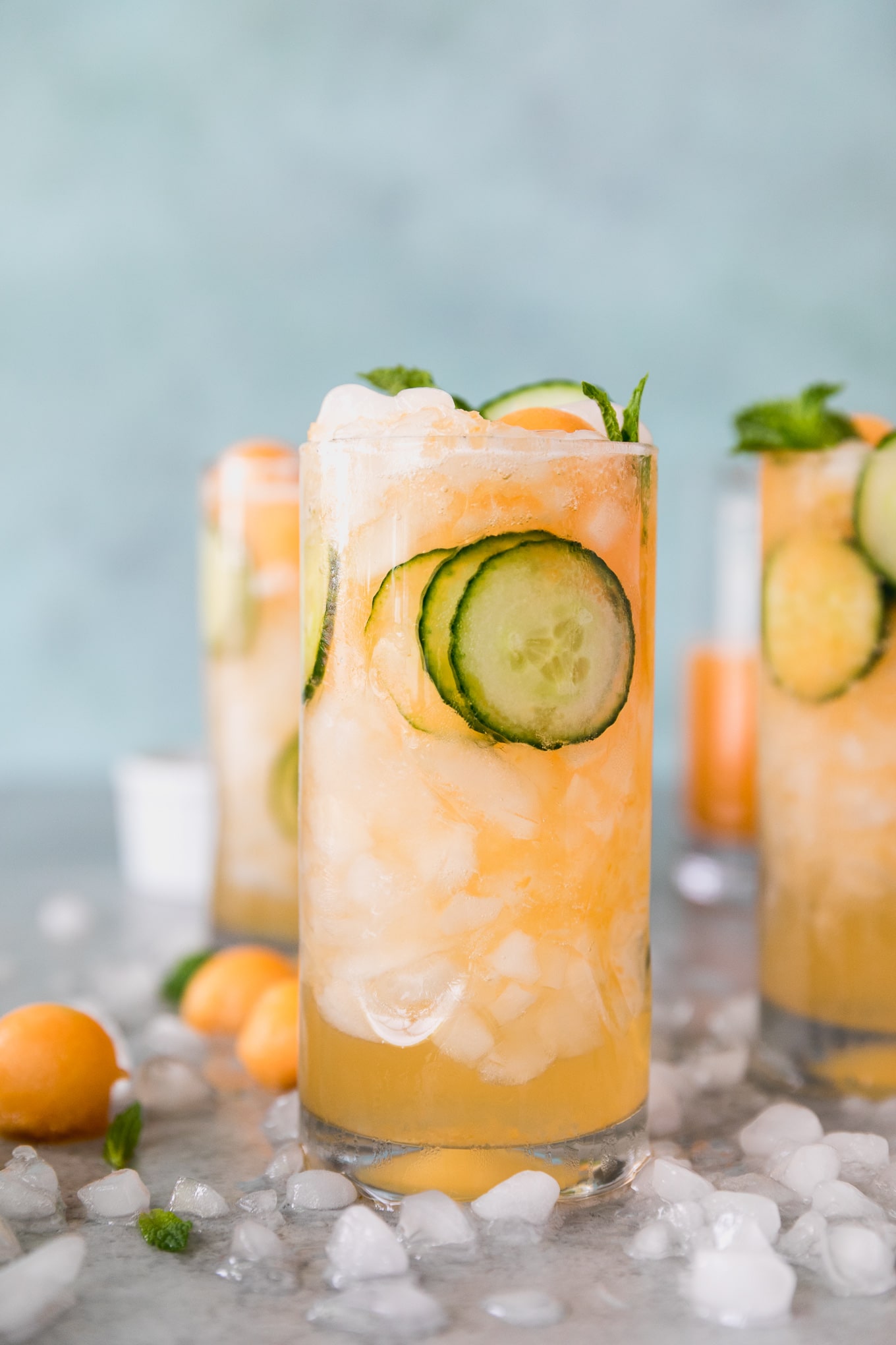 Forward facing close up shot of 3 cucumber melon cocktails with sliced cucumbers in the glasses, and salon balls and crushed ice scattered around