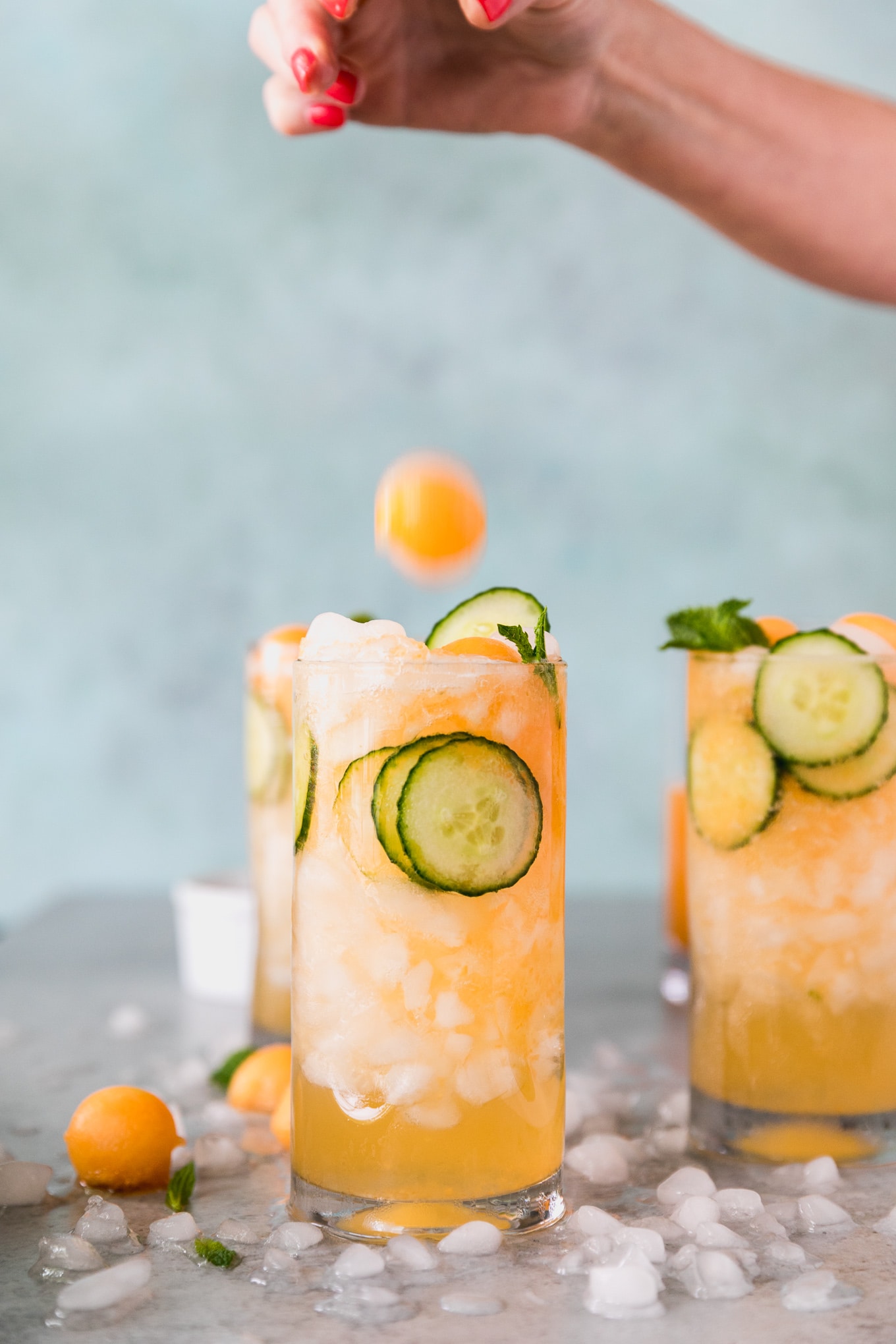 Forward facing shot of 3 cucumber melon cocktails with a hand dropping a melon ball onto the closest one in the foreground