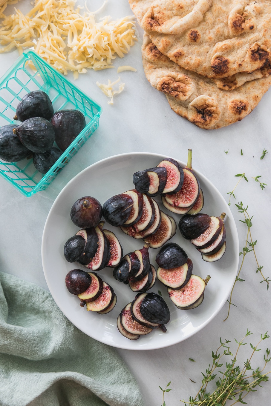 Overhead shot of a plate of sliced figs surrounded by naan bread, a turquoise basket of figs, and fresh thyme sprigs