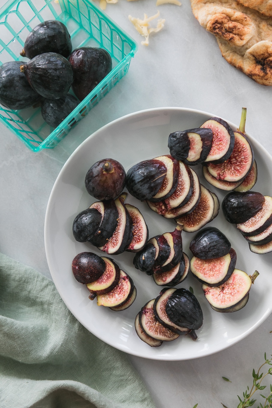 Overhead close up shot of a plate of sliced figs surrounded by a turquoise basket of figs, and fresh thyme sprigs