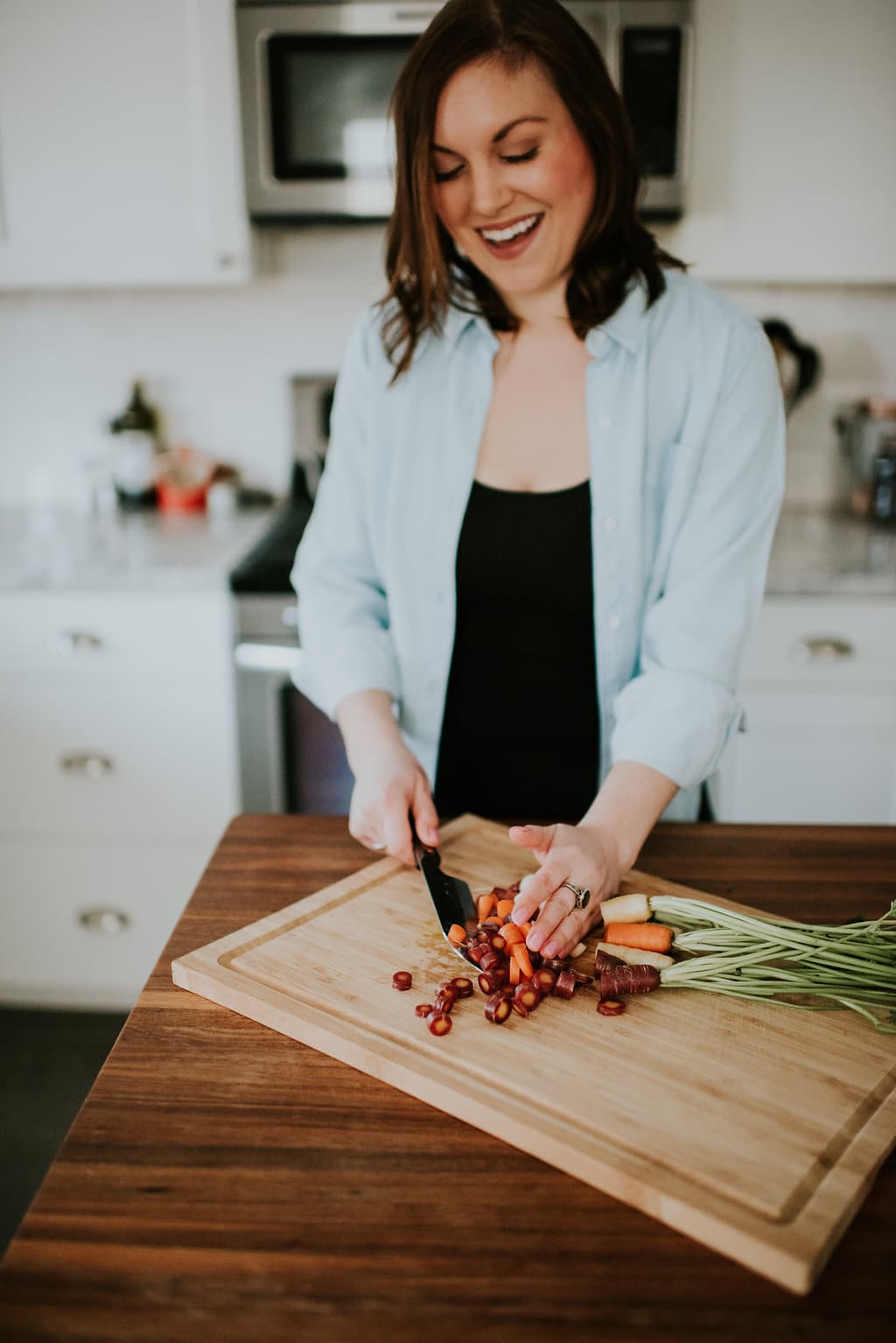 Woman smiling and laughing and scooping up chopped rainbow carrots with a chef's knife on a wooden cutting board