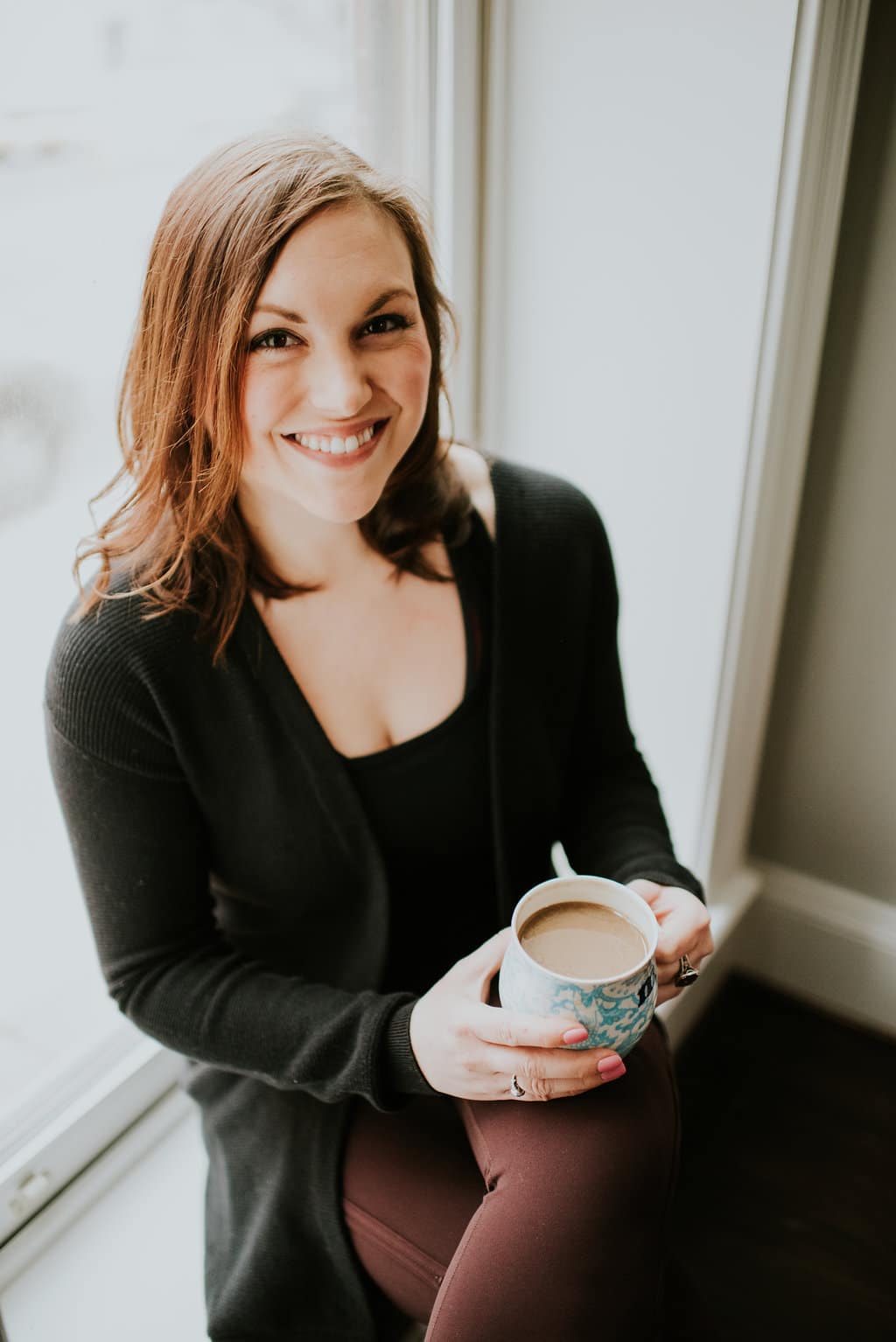 Shot of woman sitting in a windowsill smiling with a cup of coffee