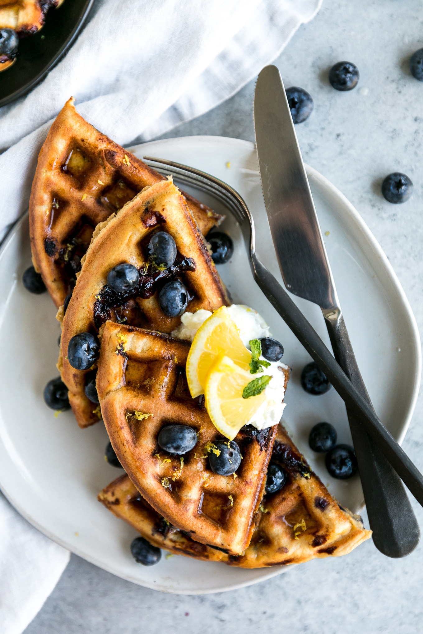 Overhead shot of a waffle cut into quarters topped with blueberries and lemon slices and a fork and knife off to the side
