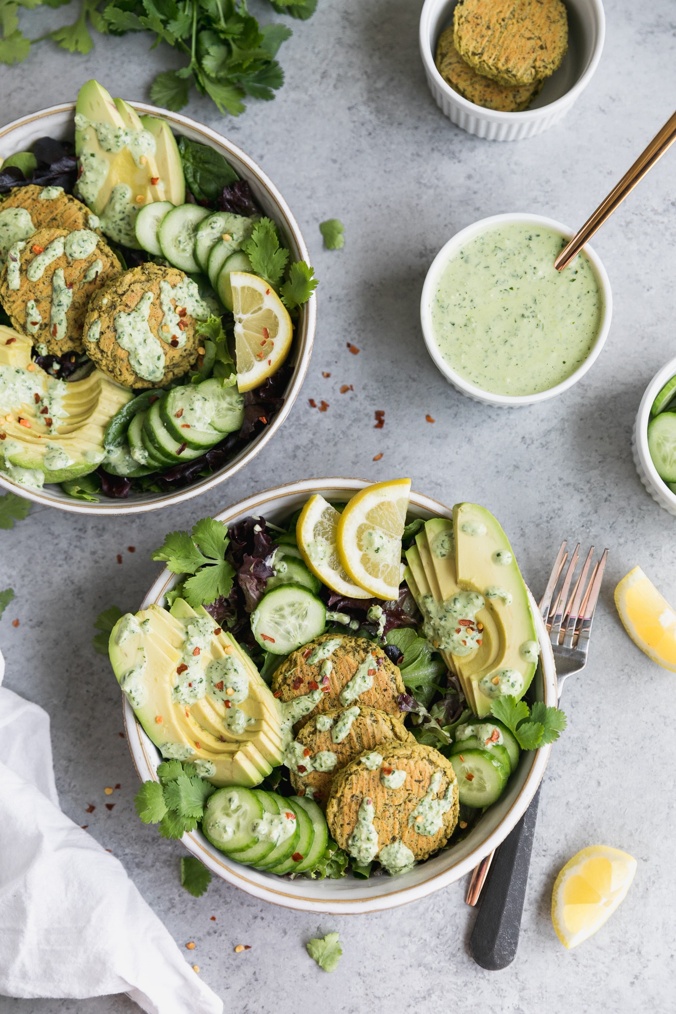 Overhead shot of two salads topped with falafel, sliced avocado, sliced cucumber, lemon slices, and yogurt sauce, with a bowl of yogurt sauce in the top right and forks resting next to the bowl