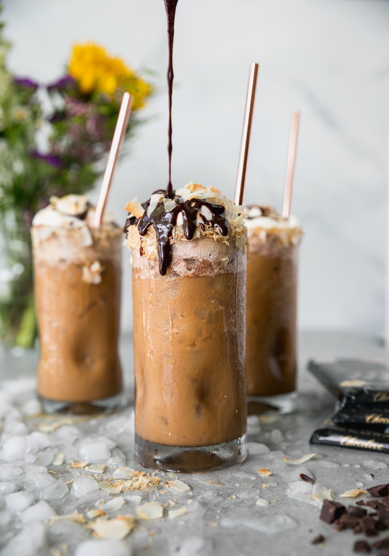 Forward facing shot of an iced coffee bring drizzled with chocolate sauce and 2 more iced coffees and a vase of flowers in the background