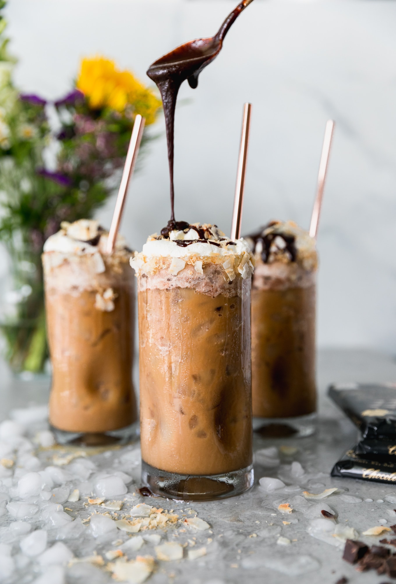 Forward facing shot of 3 iced coffees, the one in the foreground being drizzled with chocolate sauce