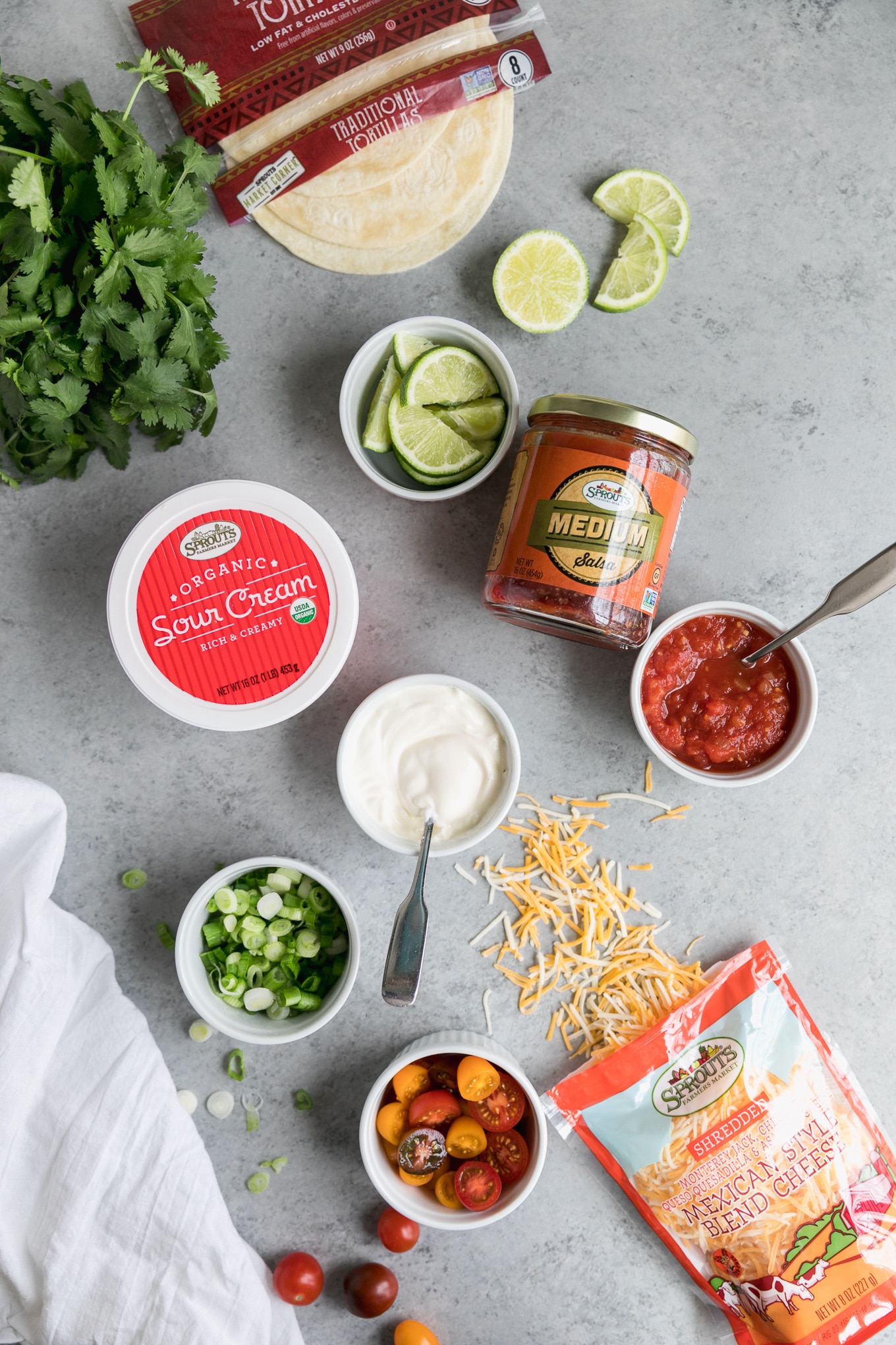 Overhead shot of small bowls filled with lime slices, sour cream, salsa, scallions, multicolored cherry tomatoes, a bag of tortillas, and a container of sour cream and a bag of cheese being spilled out in the bottom right corner