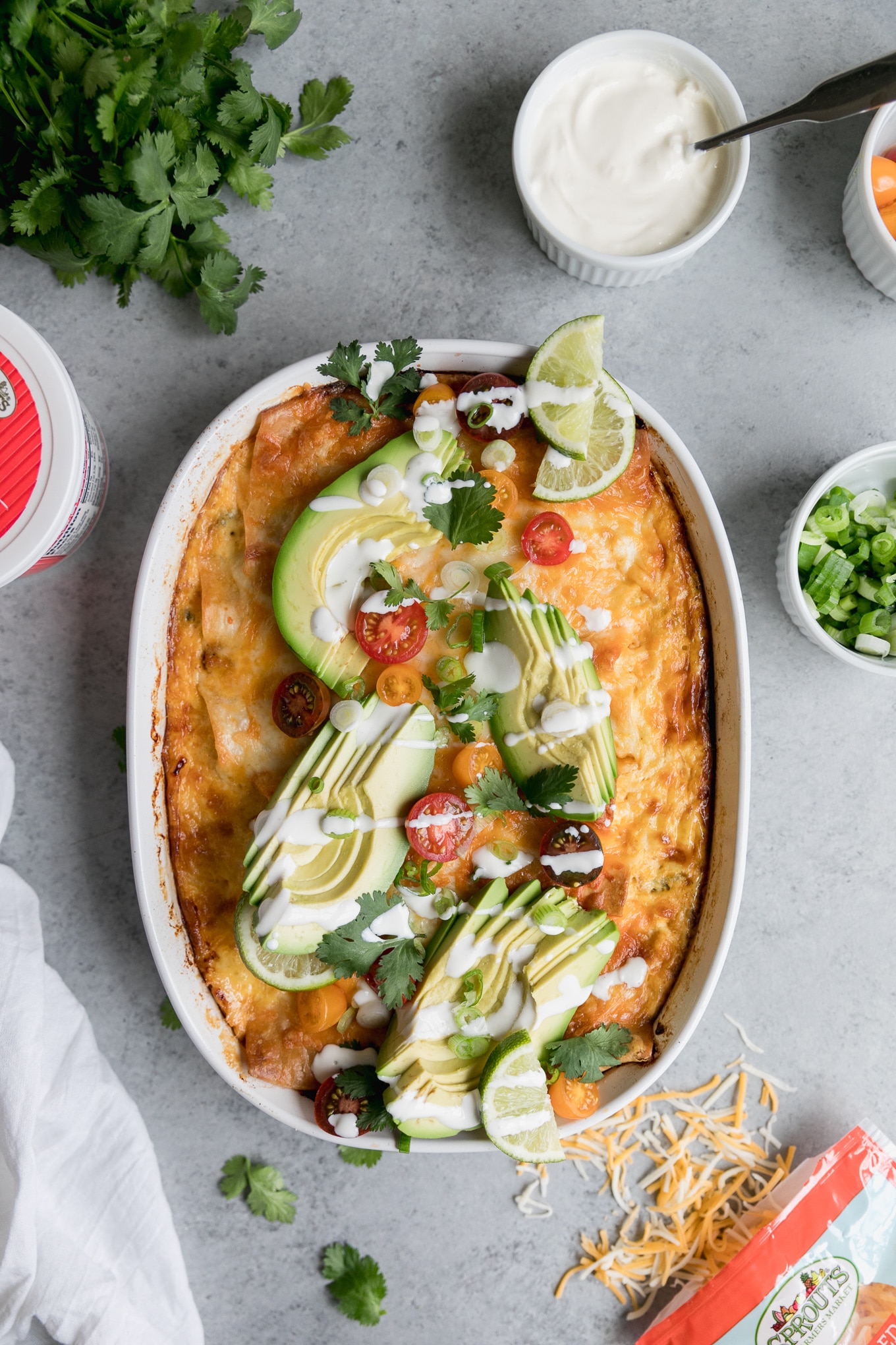 Overhead shot of a casserole dish of enchiladas topped with sliced avocado, cherry tomatoes, sur cream drizzle, and cilantro, with a container of sour cream off to the side, a bunch of cilantro in the corner, and a small bowl of scallions