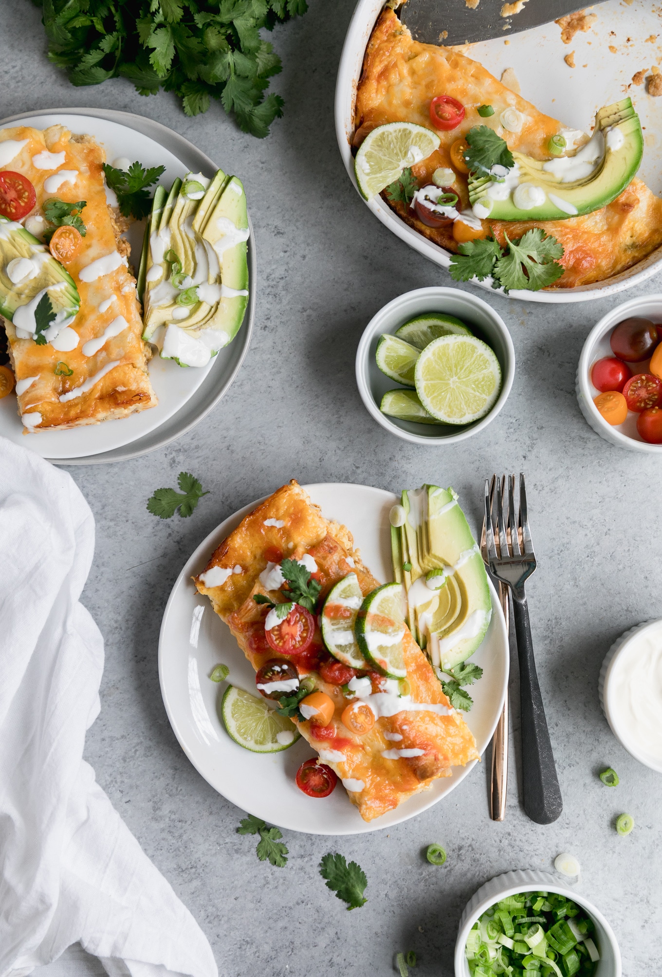 Overhead shot of two plates of enchiladas topped with halved cherry tomatoes, lime wedges, sour cream, and cilantro, with sliced avocado on the side
