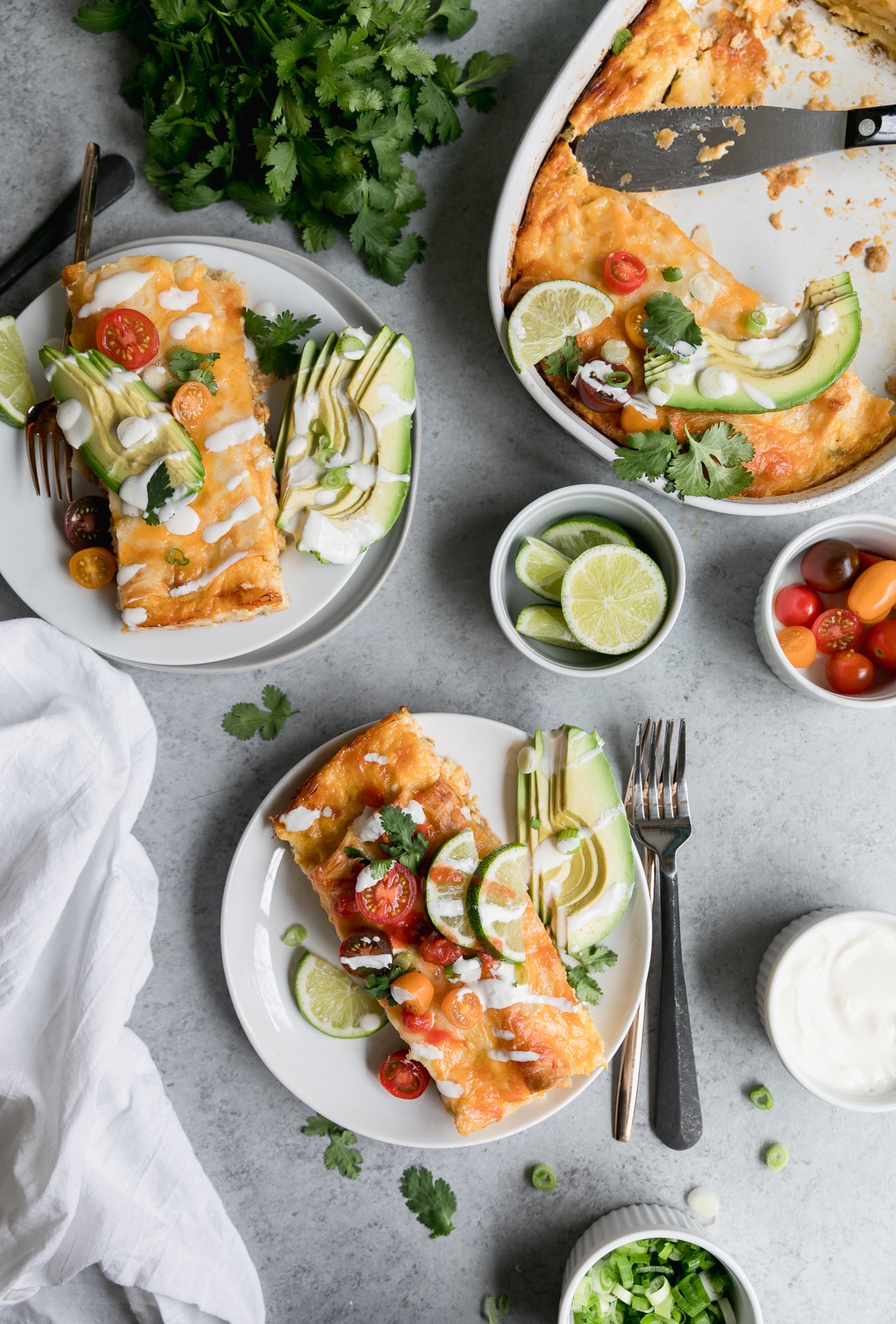 Overhead shot of two plates of enchiladas and a casserole dish of enchiladas with a bowl of cherry tomatoes, lime wedges, and sour cream off to the side
