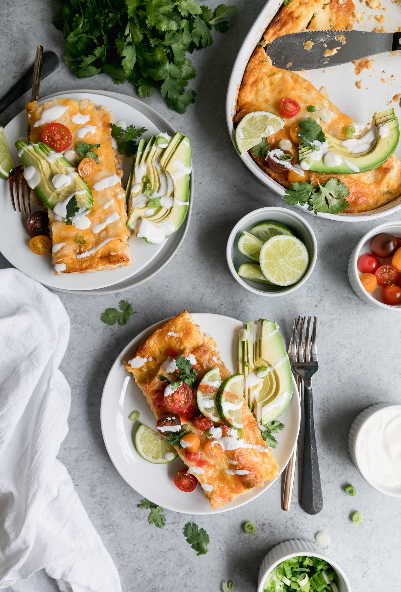 Overhead shot of two plates of enchiladas and a casserole dish of enchiladas with a bowl of cherry tomatoes, lime wedges, and sour cream off to the side