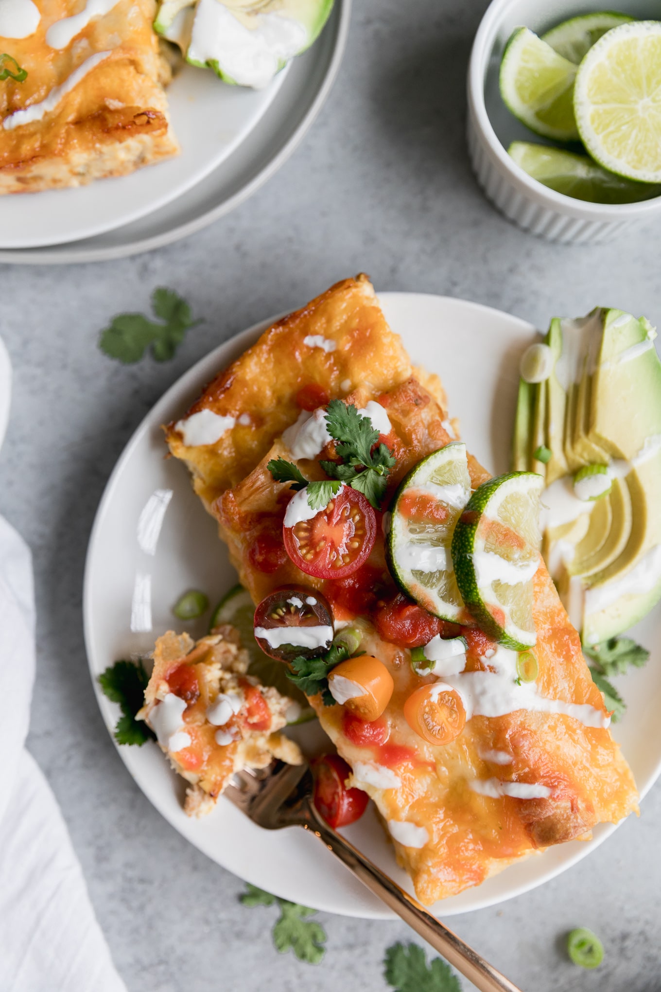 Overhead close up shot of a plate of enchiladas topped with cherry tomatoes, and lime wedges, and sour cream, with sliced avocado on the side