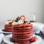 Forward facing shot of a stack of red velvet pancakes topped with whipped cream, berries, chopped dark chocolate, and mint, with another stack of pancakes behind it to the left