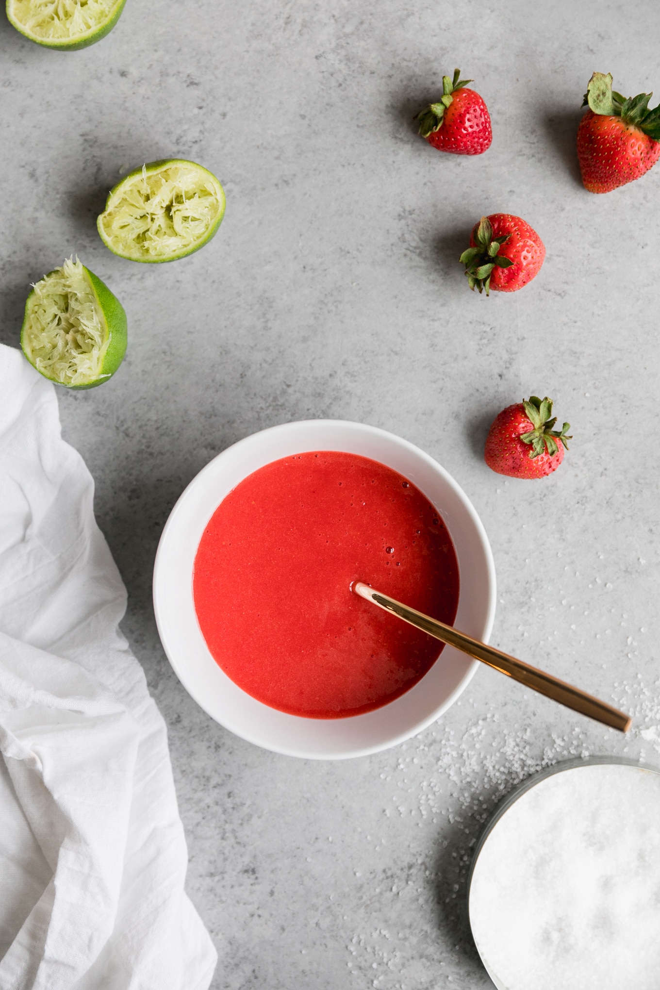 Overhead shot of a bowl of strawberry purée with a gold spoon sticking out and strawberries and squeezed limes next to it, as well as a plate of margarita salt