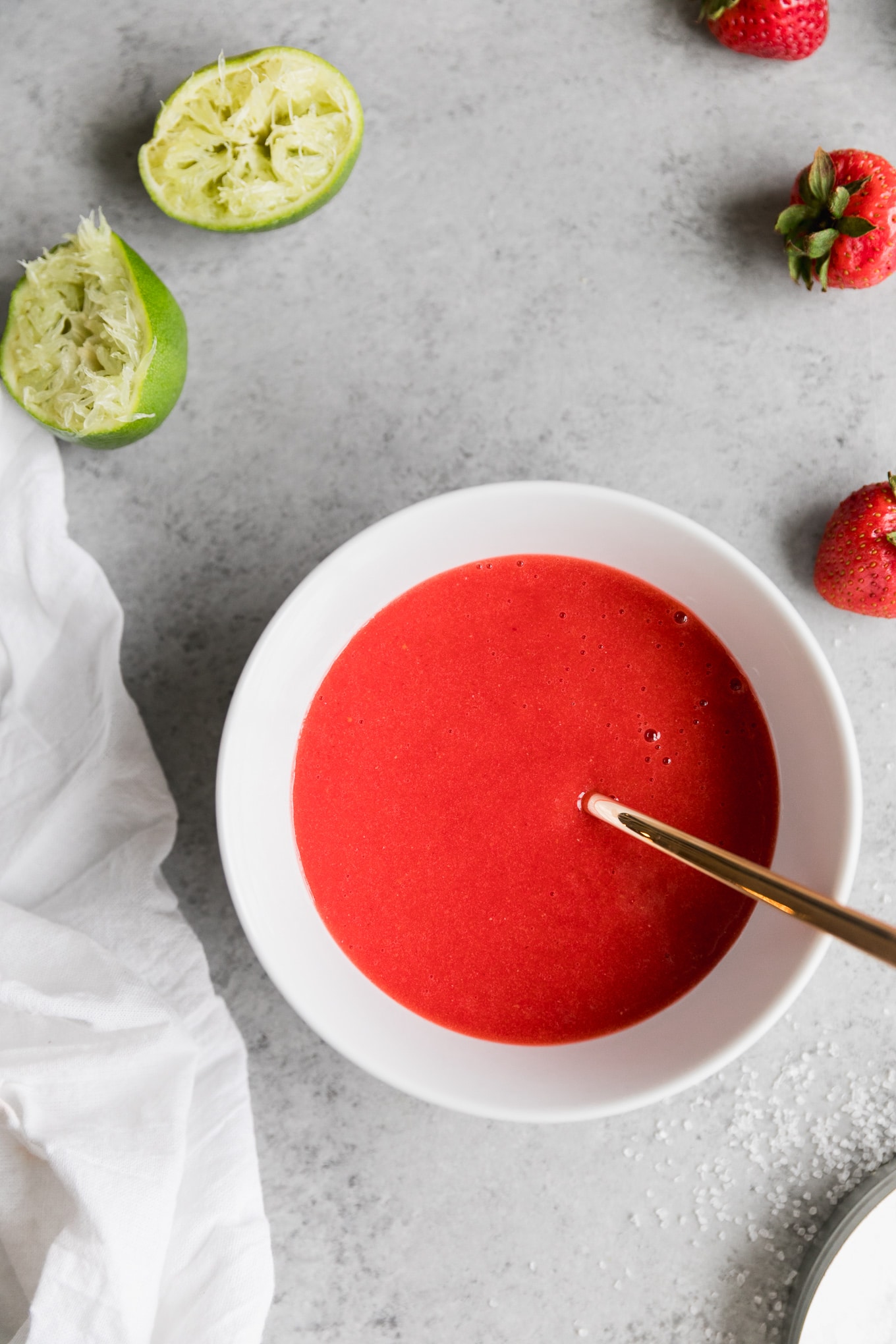 Overhead shot of a bowl of strawberry purée with a gold spoon sticking out and strawberries and squeezed limes next to it