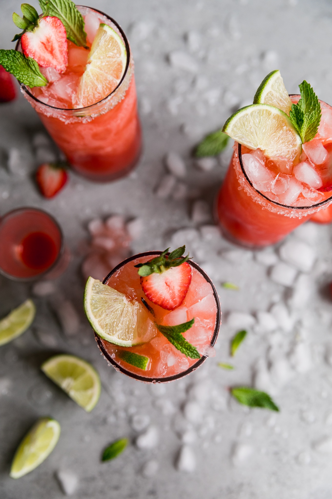 Overhead shot of three strawberry margaritas garnished with halved strawberries, mint and lime wedges, with crushed ice on the table