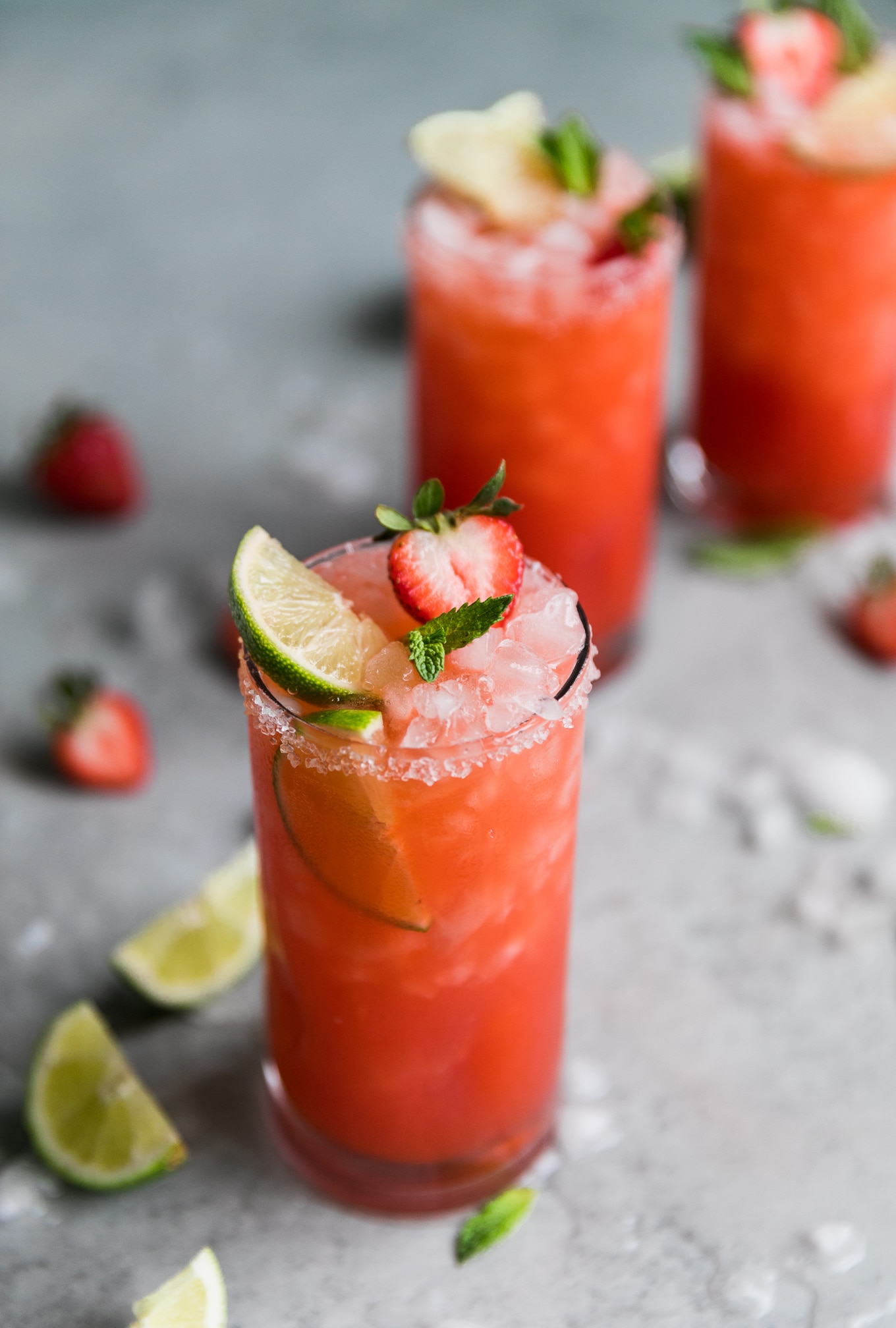 Close up shot of a strawberry margarita with a lime wedge, halved strawberry, and mint leaves sticking out, with two margaritas in the background