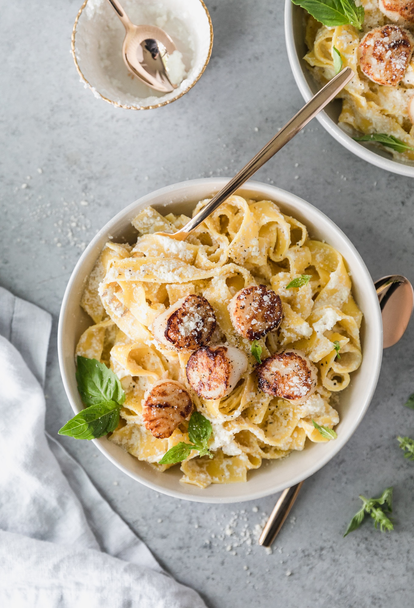Overhead shot of a bowl of pappardelle pasta topped with seared scallops, basil, and grated parmesan, with a gold fork sticking out and another bowl of pasta peeking out in the top right of the frame