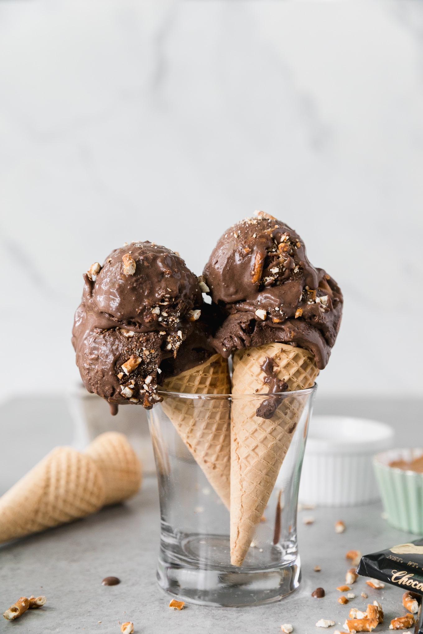 Forward facing shot of two ice cream cones with dark chocolate peanut butter pretzel ice cream in a clear glass with cones and ramekins behind it