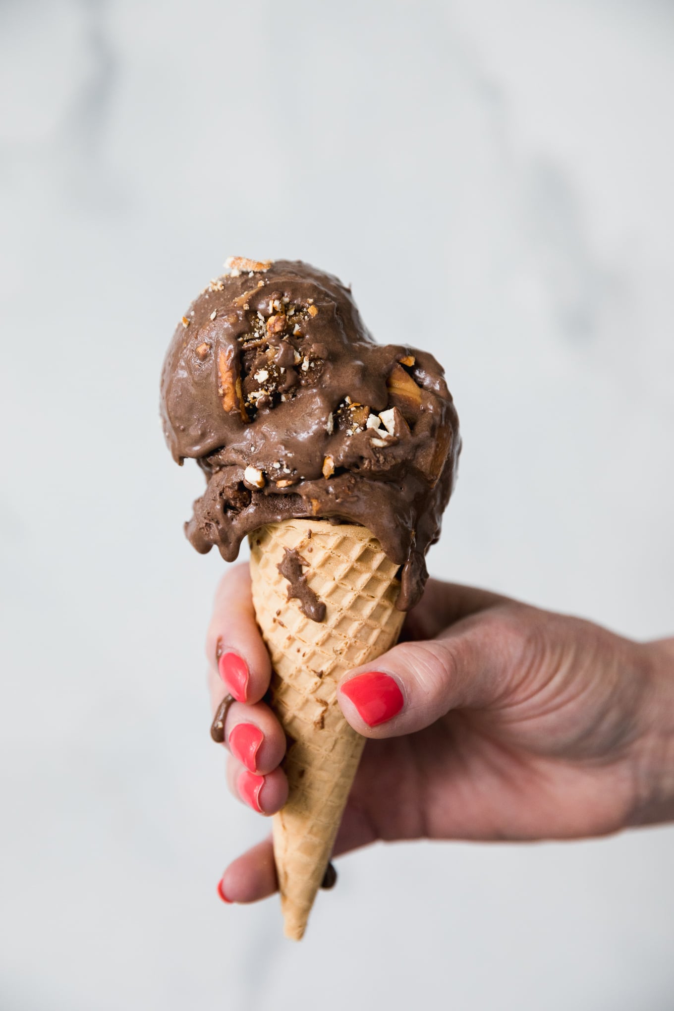 Close up forward facing shot of a hand holding a ice cream cone topped with dark chocolate peanut butter pretzel ice cream and it dripping down on the hand