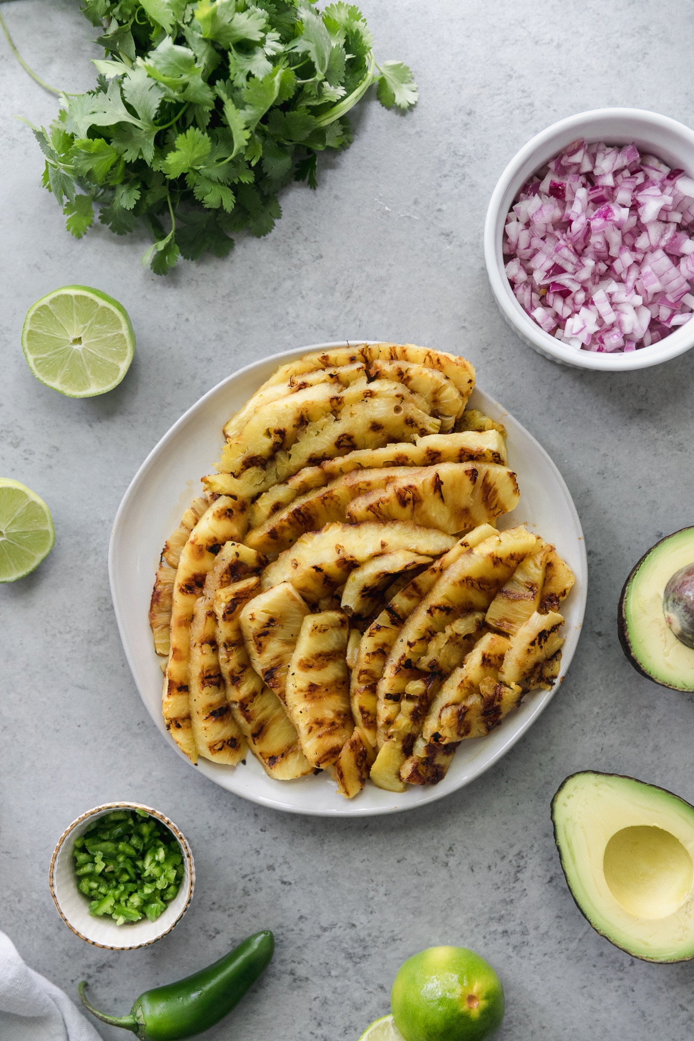 Over head shot of a plate of grilled pineapple spears, surrounded by a halved avocado, a bowl of chopped jalapeño, a bunch of cilantro, and a bowl of diced red onion