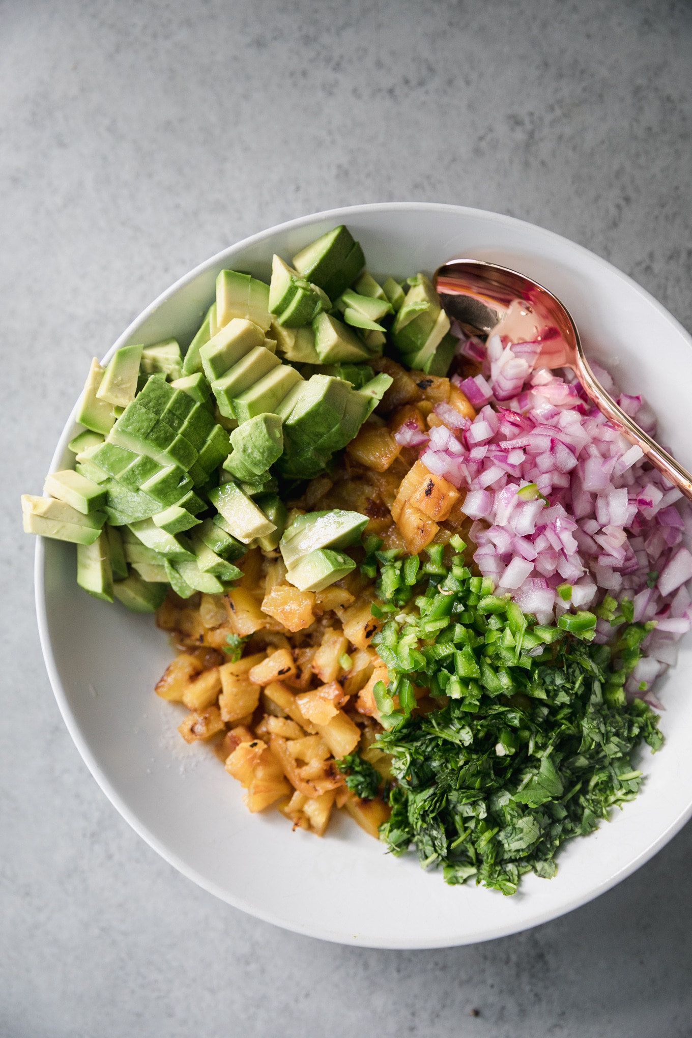 Overhead close up shot of a white bowl filled with piles of diced avocado, diced red onion, diced grilled pineapple, and chopped cilantro with a bronze spoon on the right hand side of the bowl