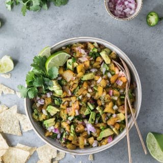 Overhead shot of a bowl of grilled pineapple avocado salsa with chips scattered to the left of the bowl, a bunch of cilantro in the top left, a bowl of diced red onion in the top right, and a squeezed lime half in the bottom right