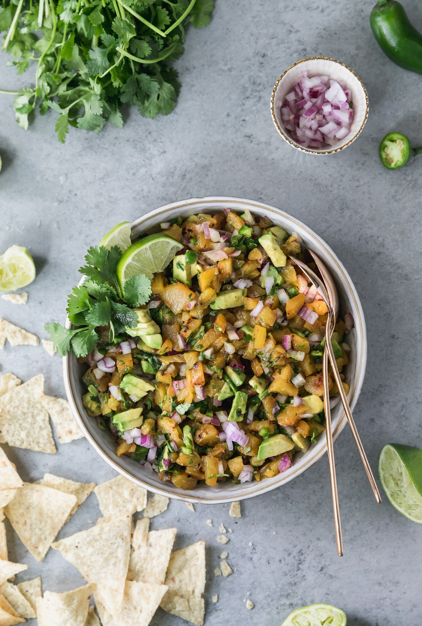 Overhead shot of a bowl of grilled pineapple avocado salsa with chips scattered to the left of the bowl, a bunch of cilantro in the top left, a bowl of diced red onion in the top right, and a squeezed lime half in the bottom right