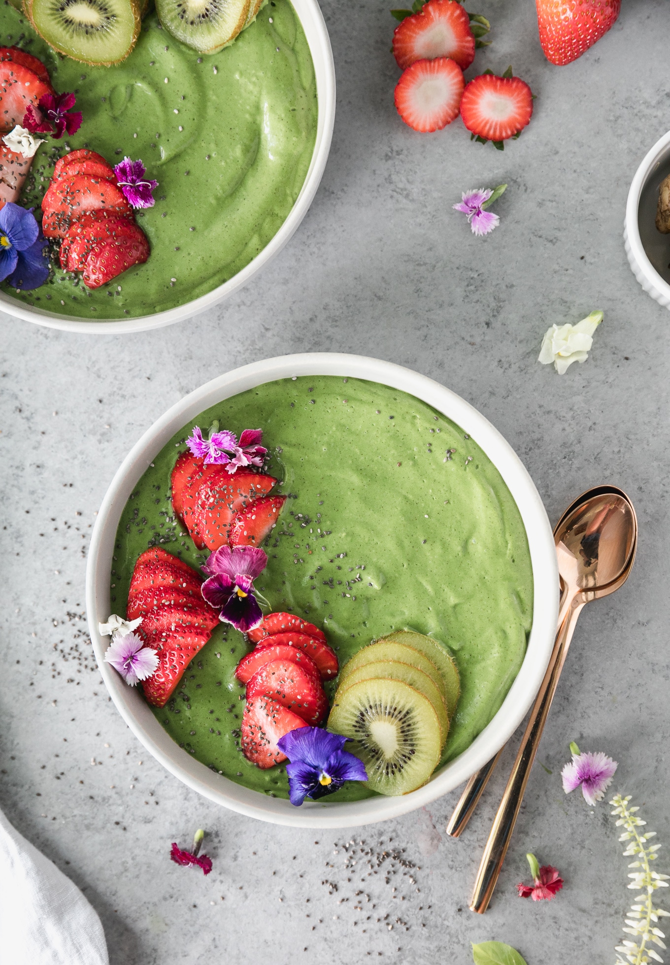 Overhead shot of a green smoothie bowl topped with sliced strawberries, kiwi, edible flowers, and chia seeds, with two gold spoons resting beside it 
