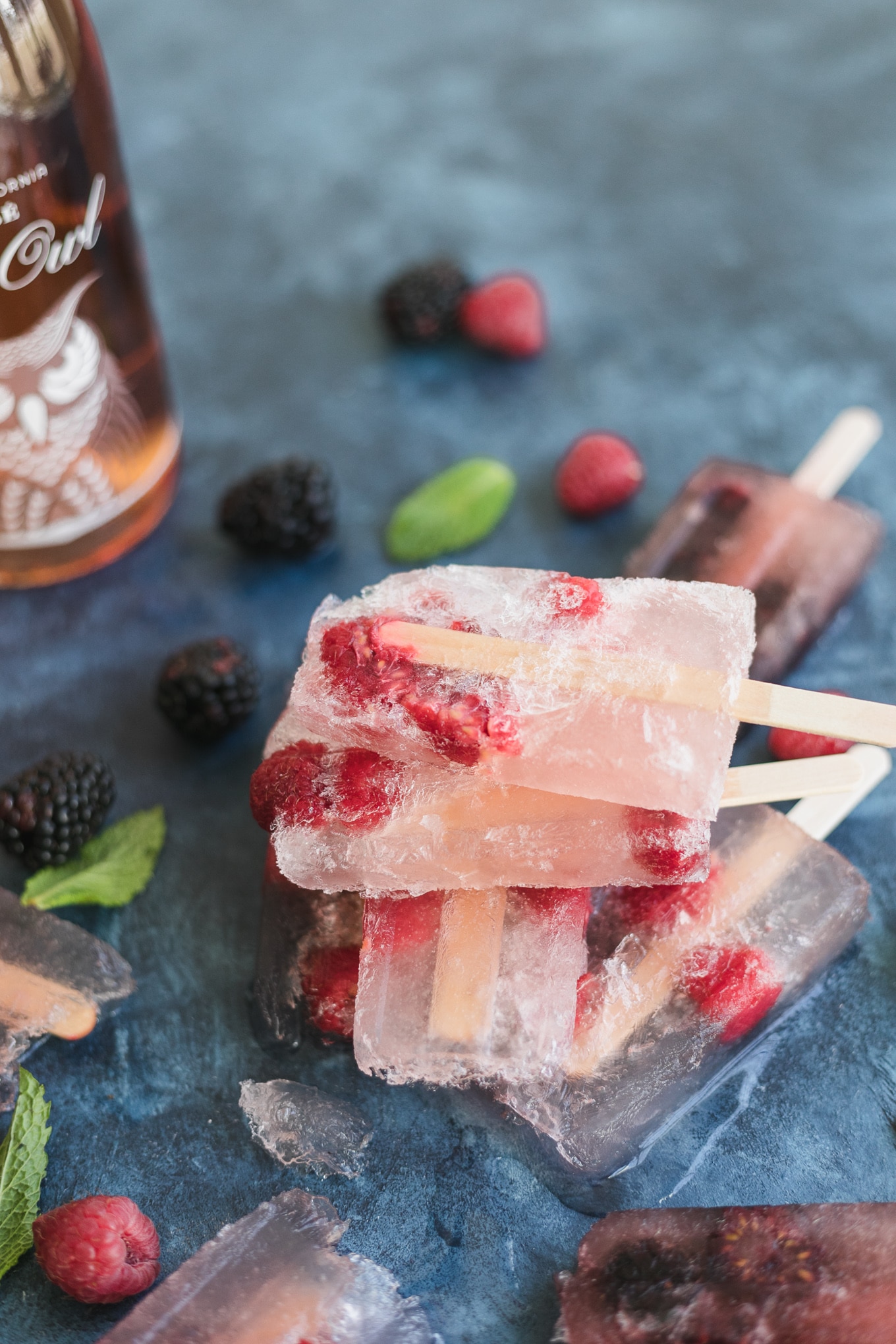 Close up shot of raspberry frosé popsicles stacked on top of each other on a blue background and berries and mint scattered around and a rosé wine bottle off to the left side
