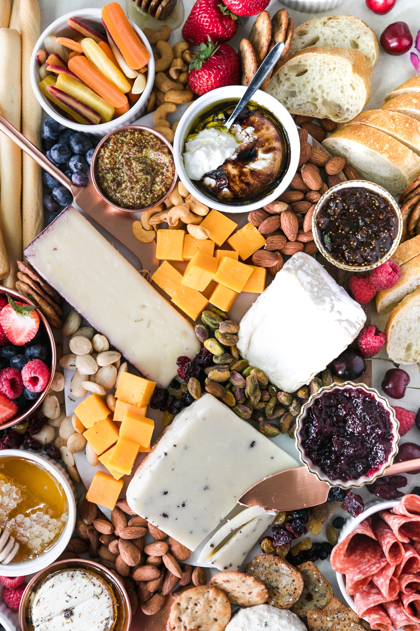 Overhead shot of a cheeseboard with various cheese, nuts, honey, preserves, honey, salami, crackers, bread, and fresh berries