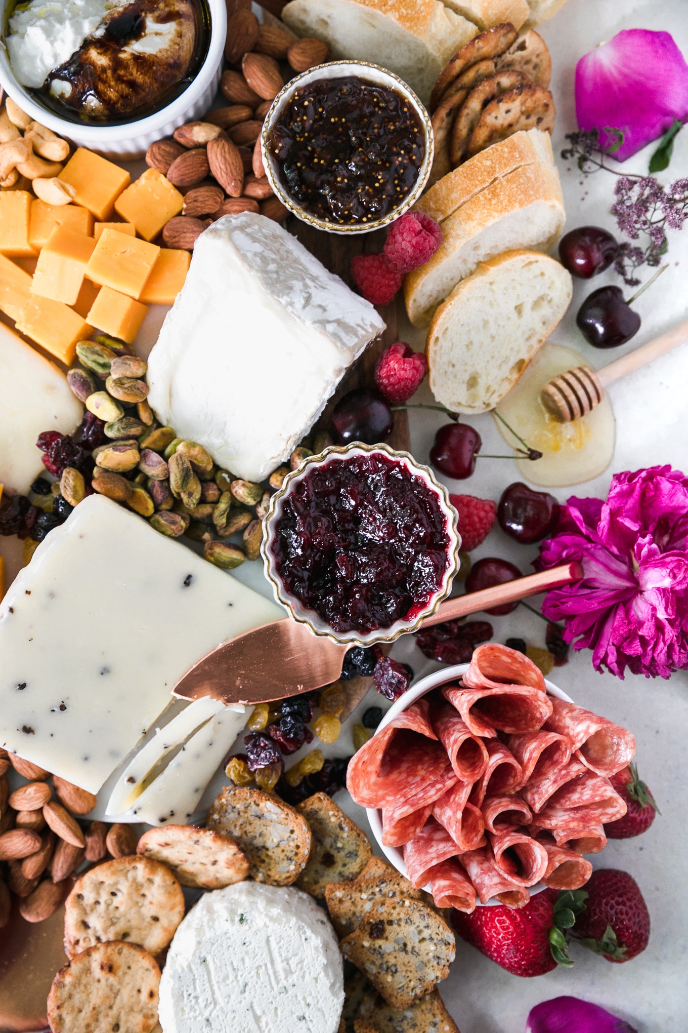 Overhead shot of a cheeseboard with various cheese, nuts, honey, preserves, honey, salami, crackers, bread, and fresh berries and a pink peony flower off to the side