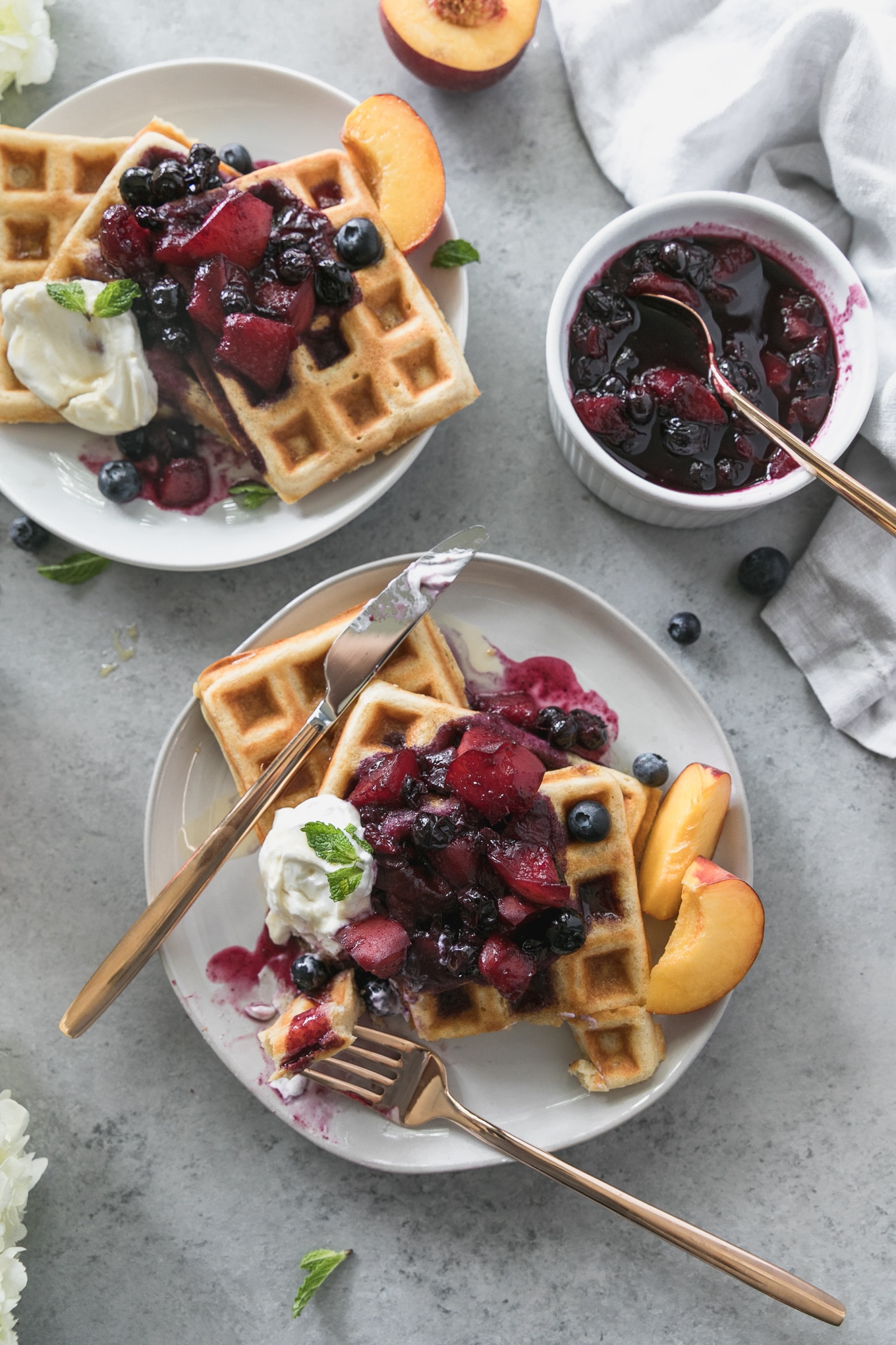 Overhead shot of two plates of waffles with peach blueberry compote on top, with a bite taken out of the waffles on the lower plate and a bowl of compote in the top right