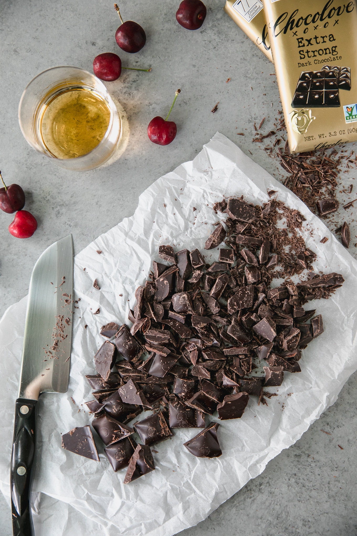 Overhead shot of a pile of chopped dark chocolate with a knife next to it as well as fresh cherries, a glass of bourbon, and a dark chocolate ba