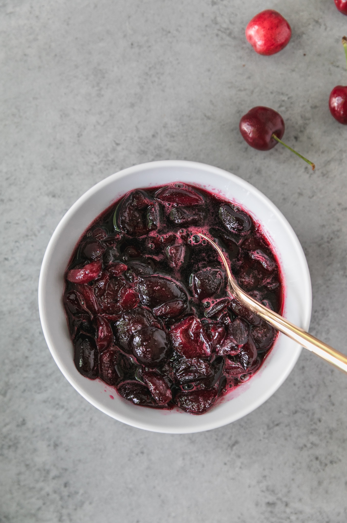 Overhead shot of a white bowl filled with bourbon macerated cherries and a gold spoon in the bowl
