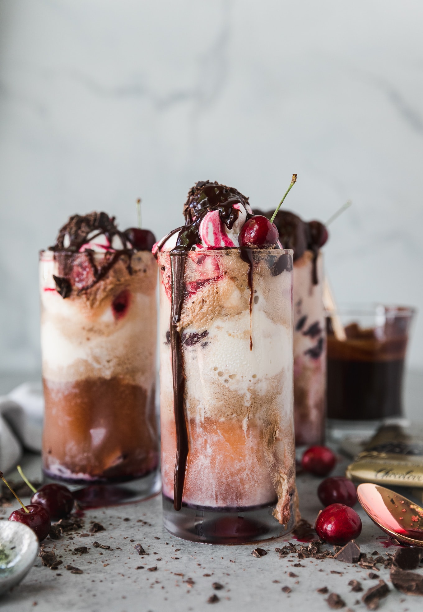 Forward facing shot of a chocolate cherry bourbon float garnished with chocolate fudge sauce, whipped cream, and cherries with two more floats behind it and a glass of chocolate fudge sauce