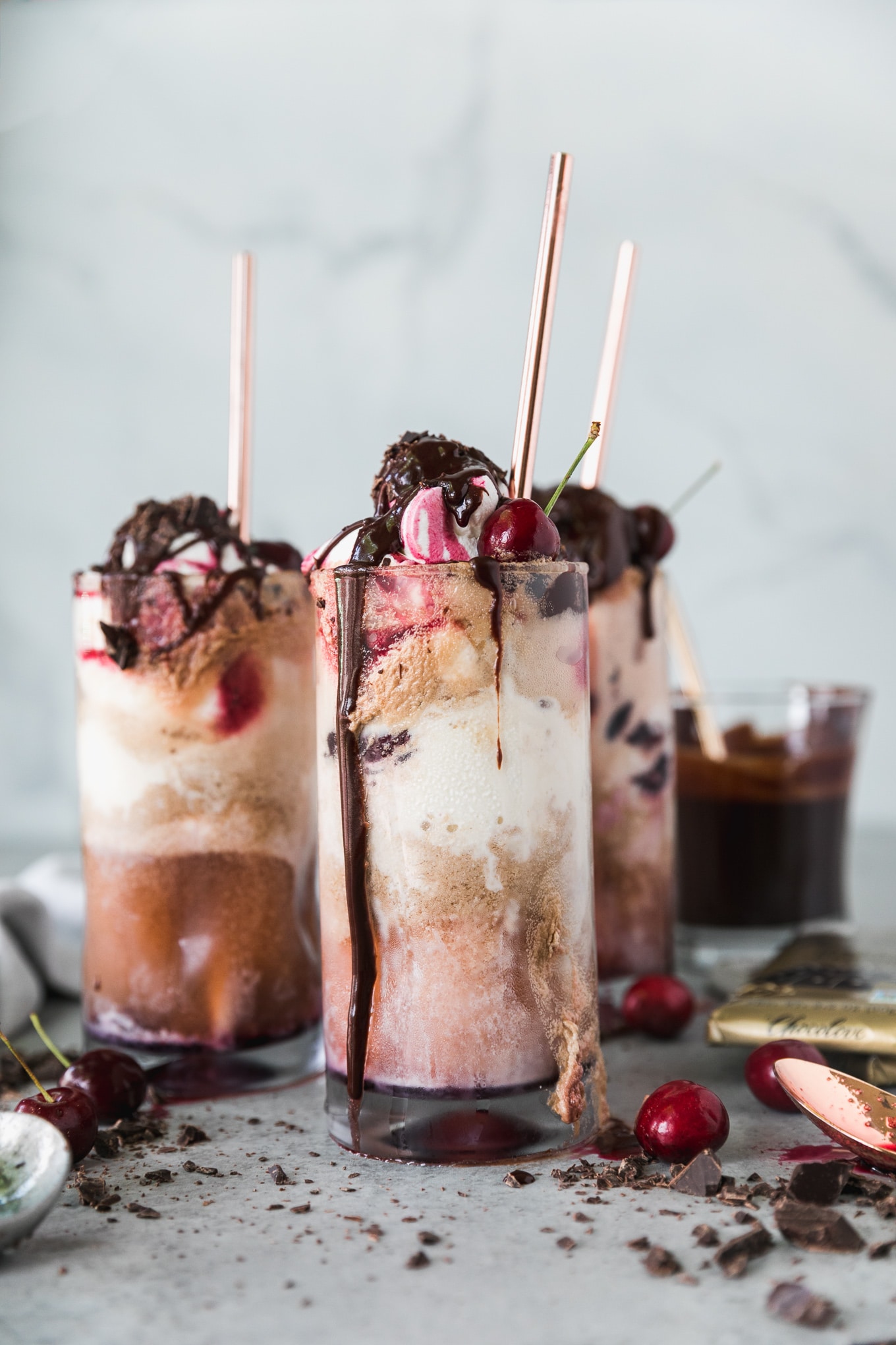 Forward facing close up shot of a chocolate cherry bourbon float topped with whipped cream and cherries, with chocolate sauce dripping down the glass and two more glasses behind it with bronze straws