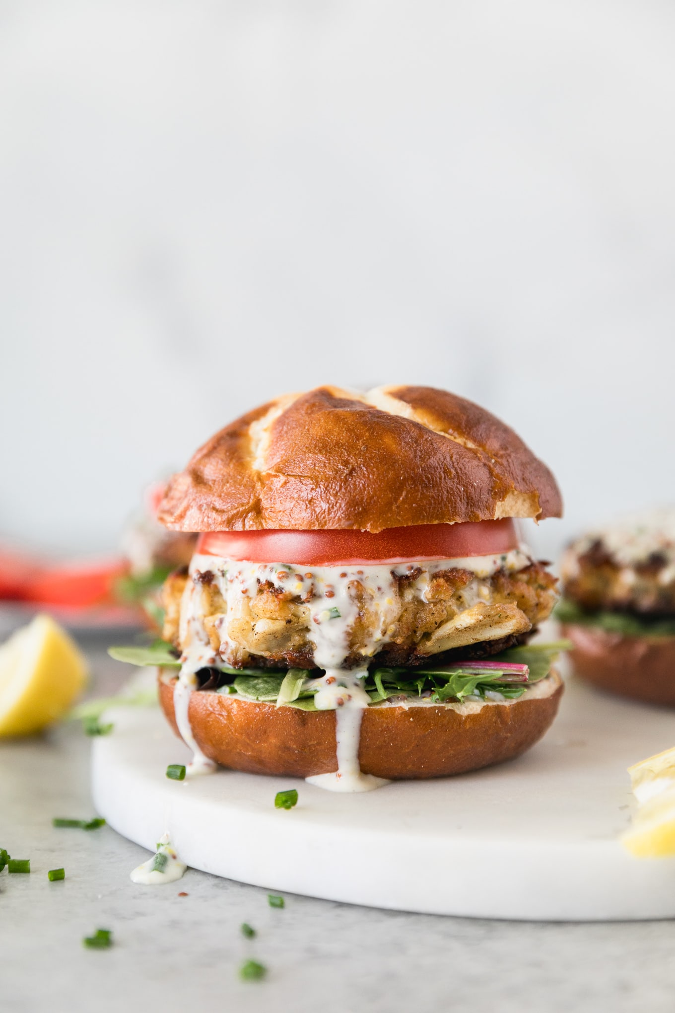 Forward facing close up shot of a crab cake sandwich with lettuce, tomato, and chive honey mustard sitting on a marble board with lemon wedges next to it