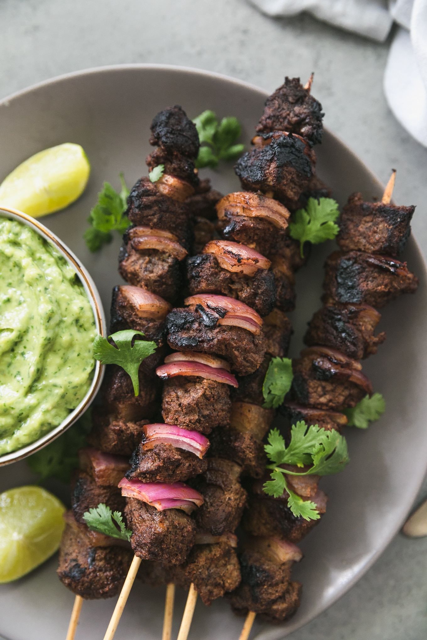 Overhead close up shot of a plate with steak and red onion skewers on it sprinkled with cilantro and a bowl of green avocado salsa on the left