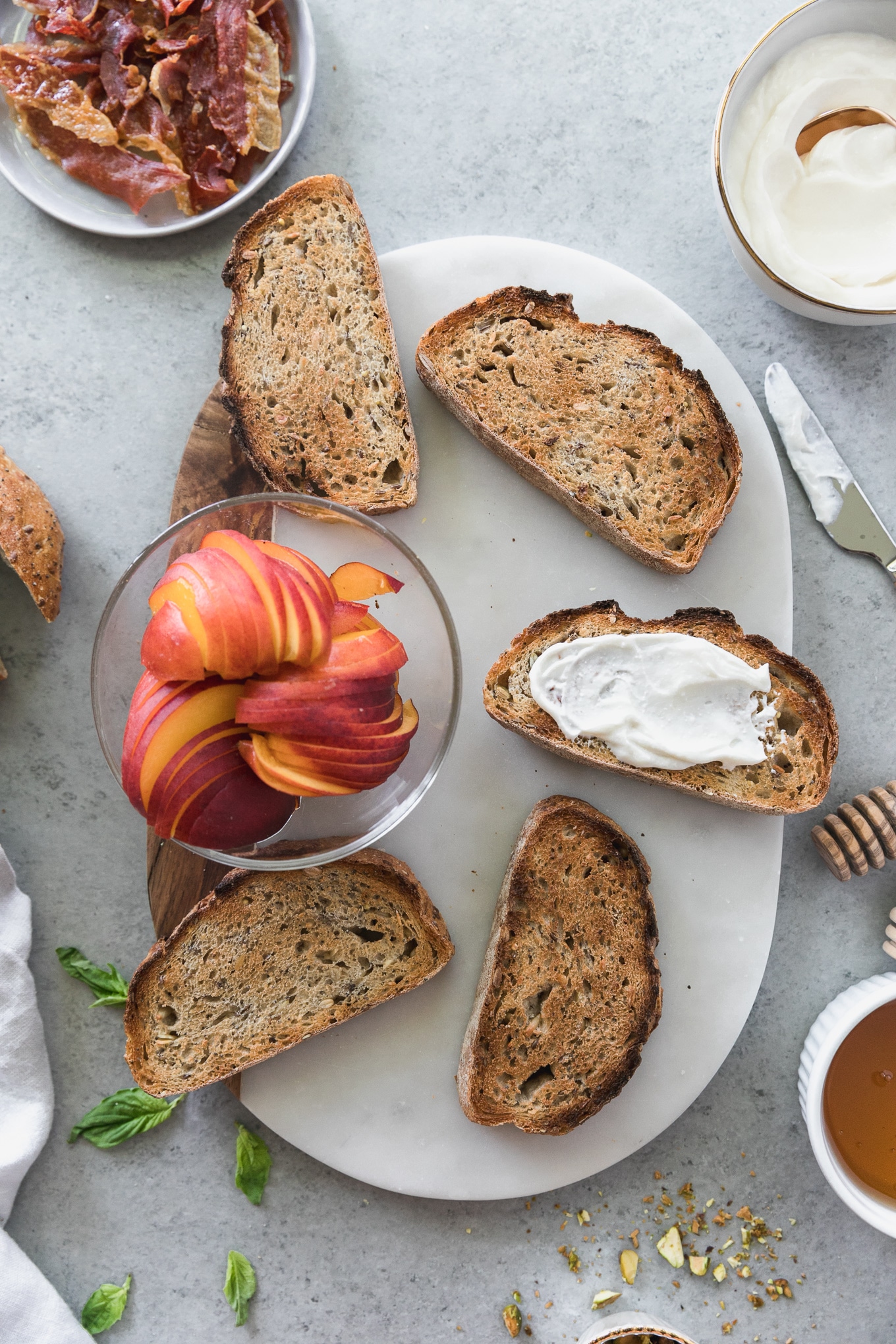 Overhead shot of a board topped with 5 slices of toast and a bowl of sliced peaches with a honey dipper, knife, bowl of whipped ricotta, and bowl of honey next to it